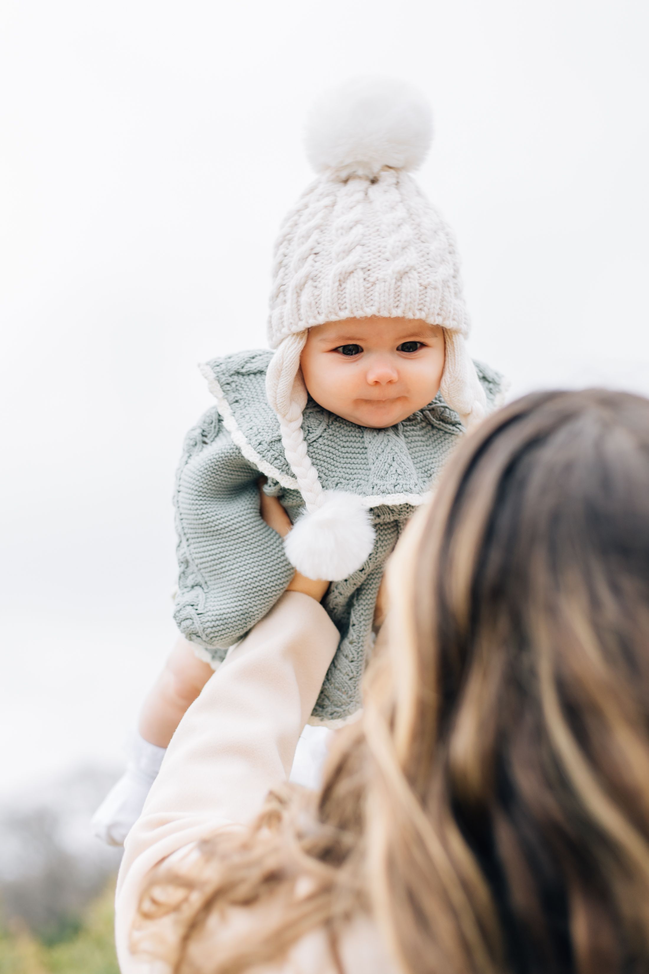 A baby wearing a white knit hat and mint green sweater being held up against a bright white background.