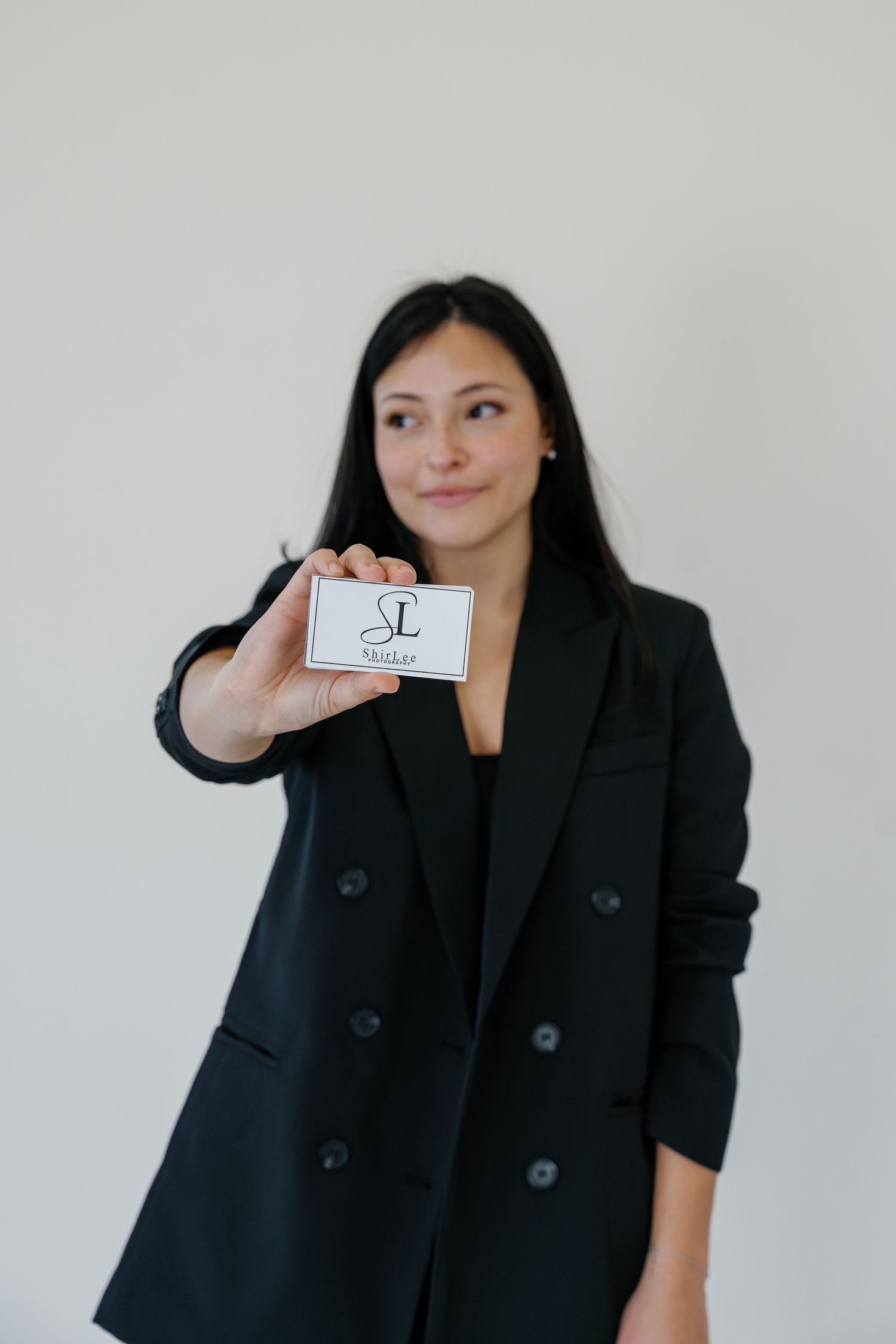 Professional standing against a light wall holds up a business card while wearing a stylish black blazer.