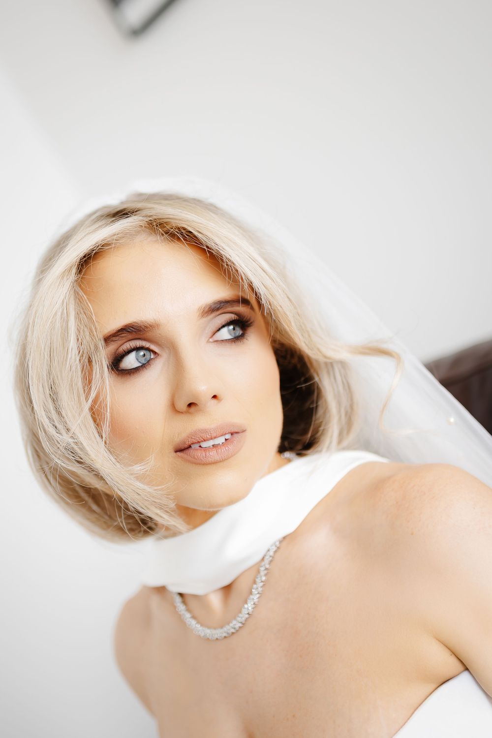 Blonde bride with blue eyes wearing a diamond necklace and white veil, elegant bridal makeup.