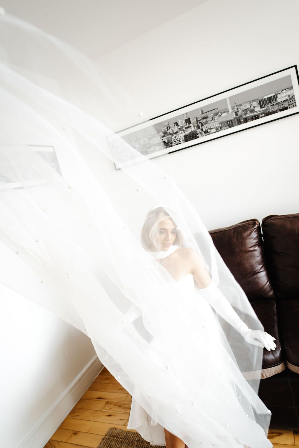 Bride in white wedding dress and veil floating in elegant room with leather sofa and black and white artwork.