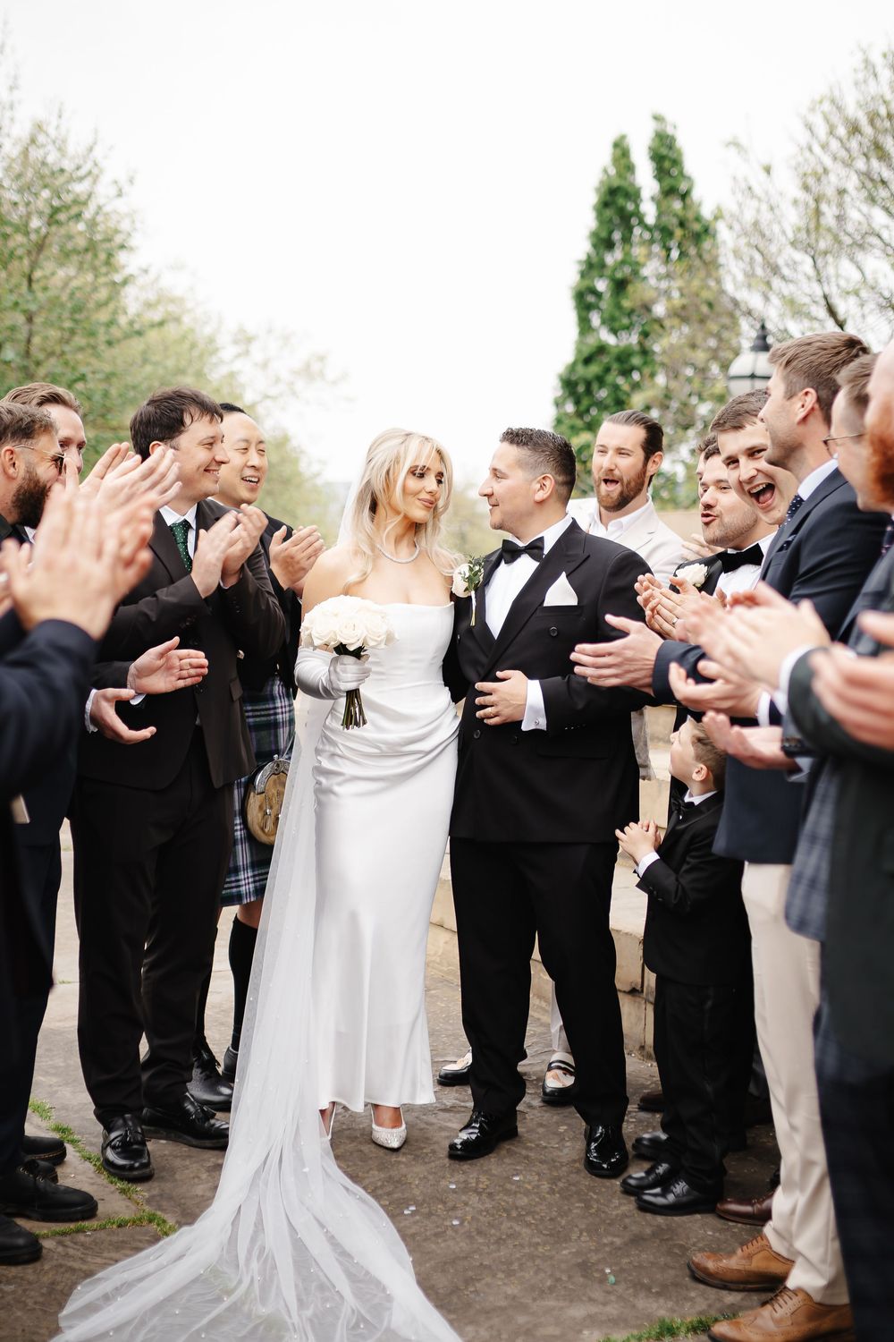 Bride in white gown and groom in tuxedo walk through cheering wedding guests outdoors.