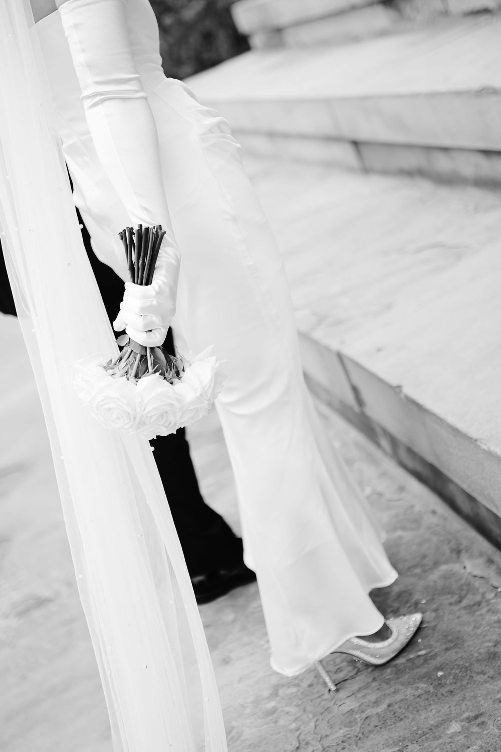 Black and white photo of bride in white gown holding bouquet with veil flowing outdoors.
