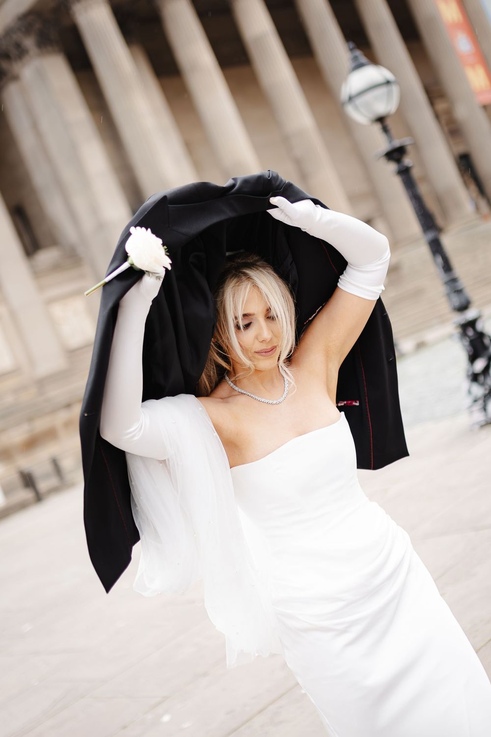 Blonde bride in white strapless gown and gloves holding black jacket overhead outdoors.