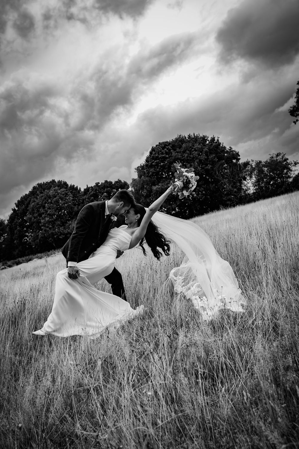A flowing white dress billows dramatically across a grassy hillside field under moody storm clouds in black and white.