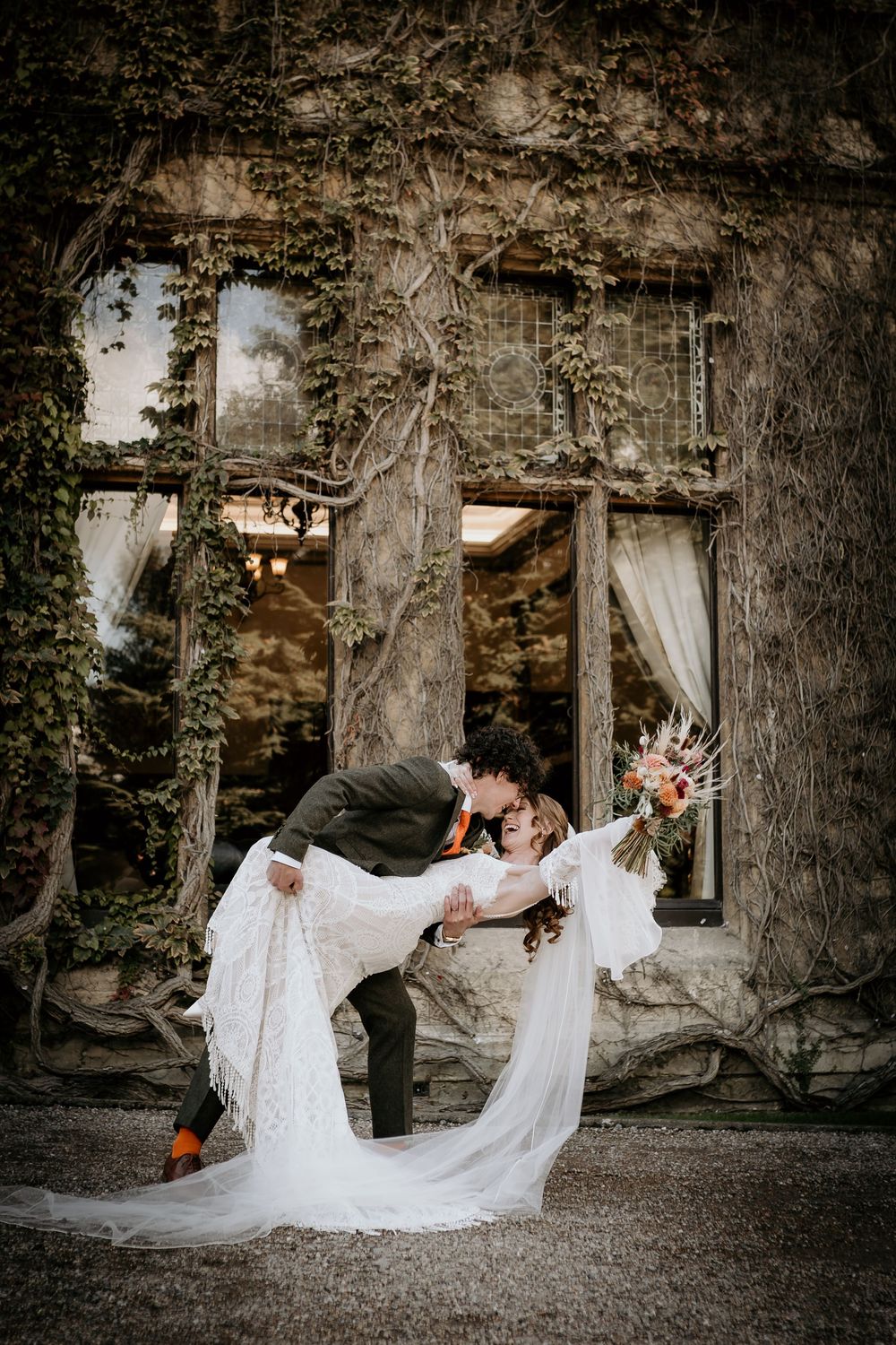 A romantic wedding photo in front of an ivy-covered stone wall with flowing white dress and dramatic dip pose.