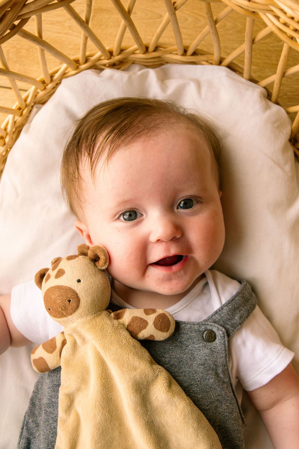 Infant lying on a wicker chair with a plush giraffe lovey wearing a gray outfit.