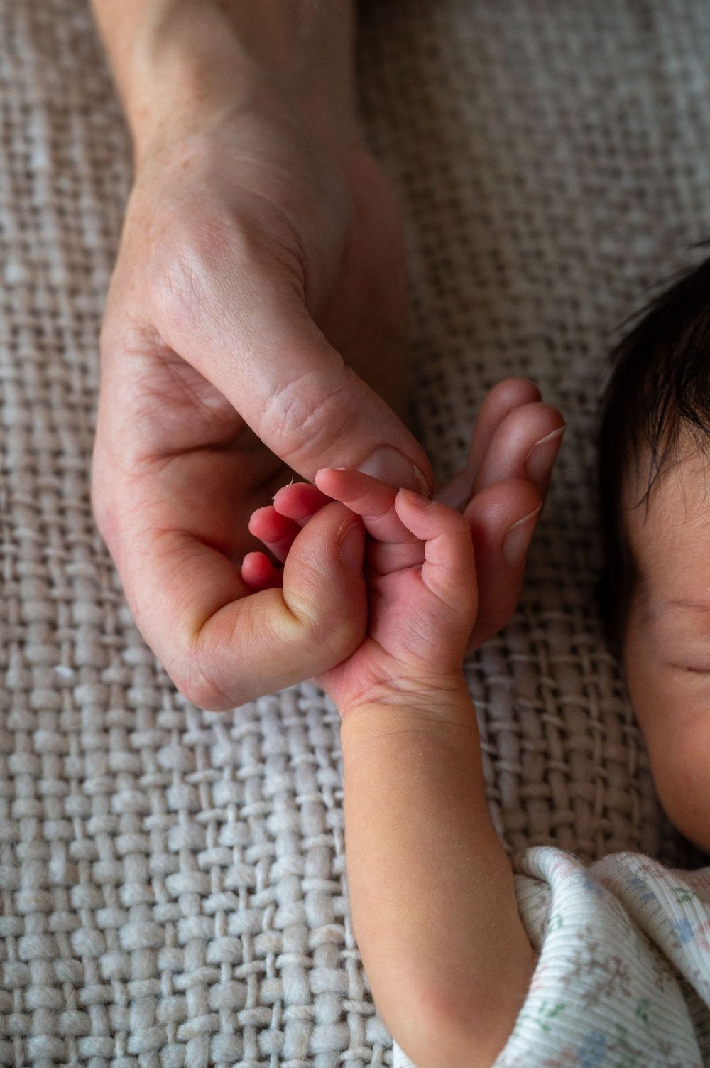 Tender moment showing mother's hand gently holding tiny newborn baby's fingers against textured fabric.