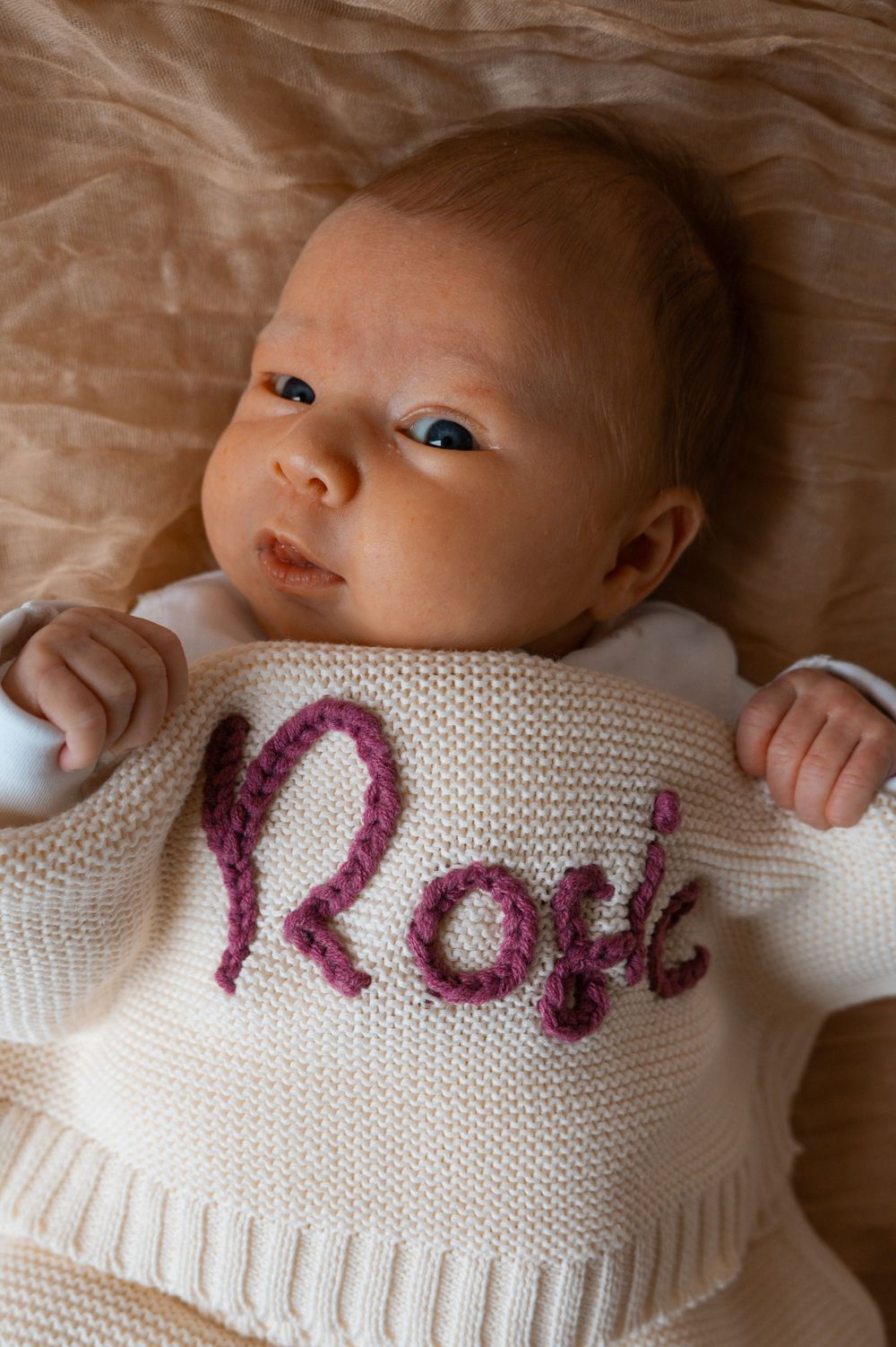 A newborn baby in white knit clothing with pink lettering while lying on beige bedding.