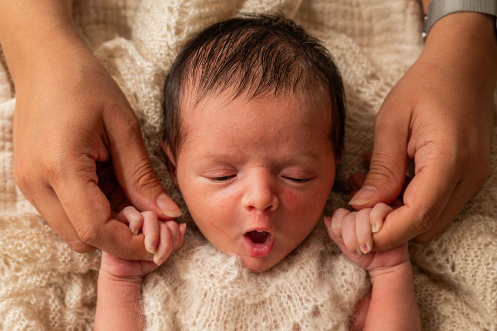 A sleeping newborn baby's face framed by mother's hands against a cream knit background.