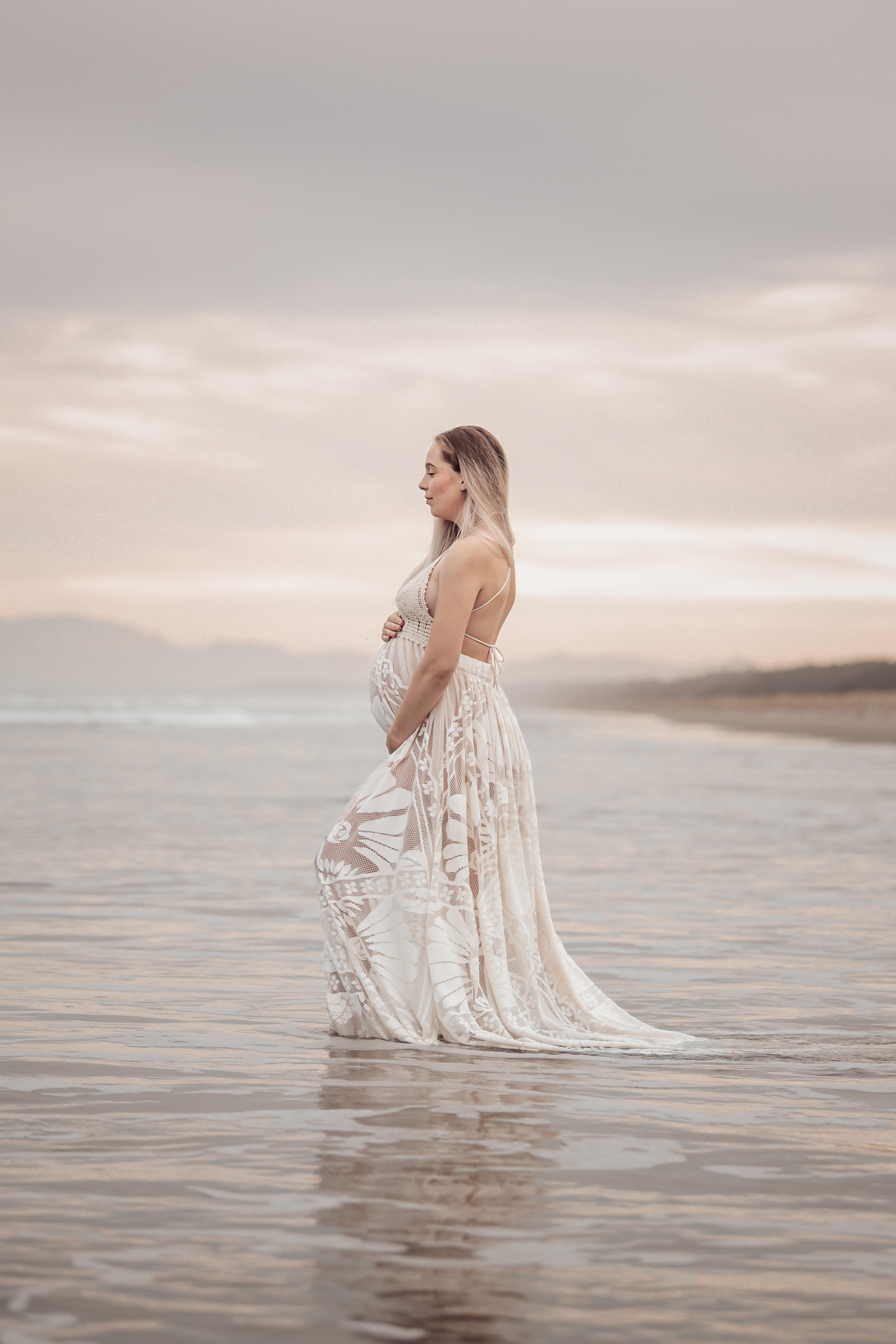 Elegant maternity photo with woman in lace dress standing in calm ocean waters at dusk.