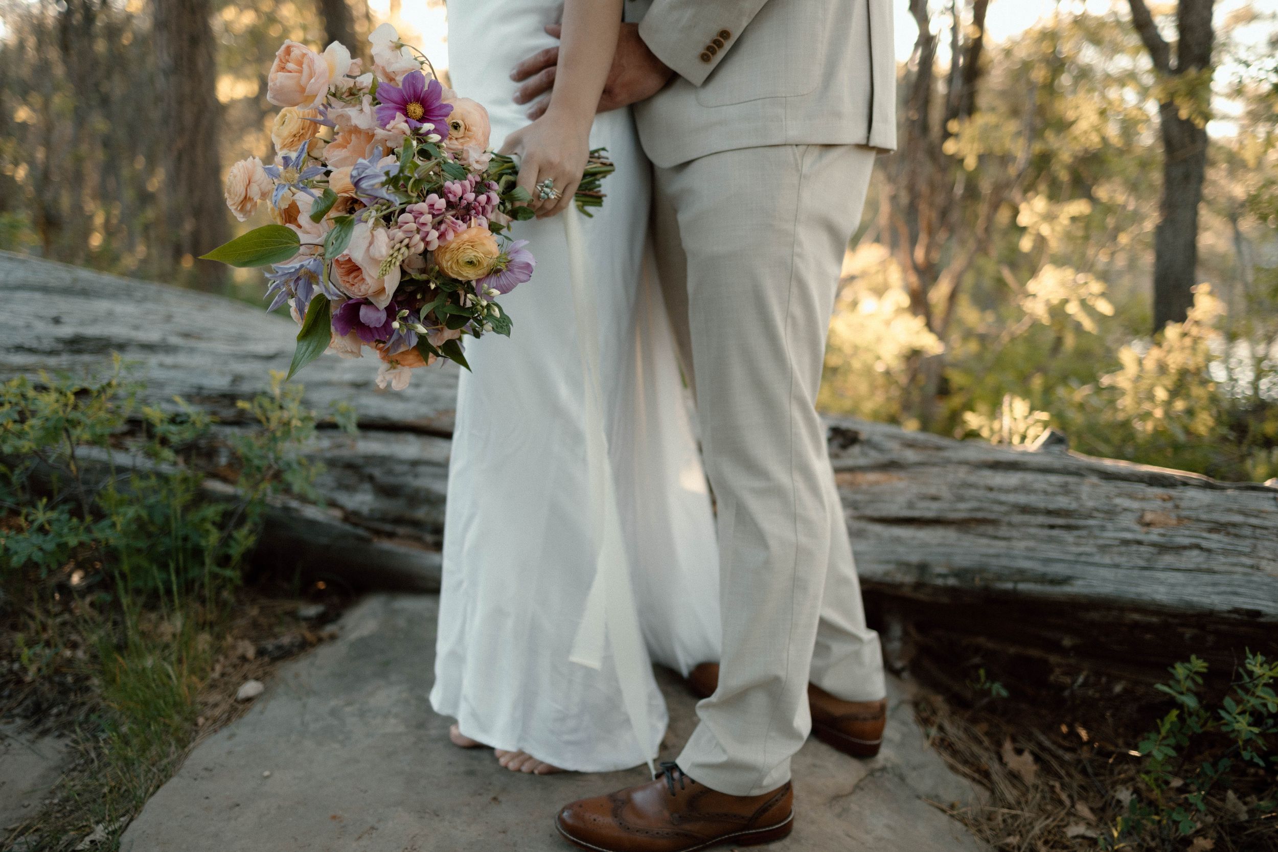 Couple in white wedding attire holding colorful bouquet on rustic wooden surface.