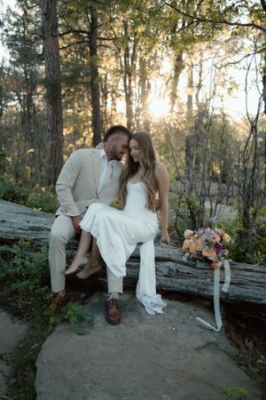 A couple in formal wear sits together on a fallen log in a sunlit forest.