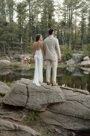 Wedding couple standing on rock overlooking serene lake surrounded by pine forest.