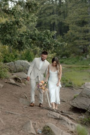 A couple in formal attire walks hand in hand down a rocky forest trail.