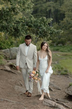 A couple walks along a rocky path holding a peach and pink floral bouquet.