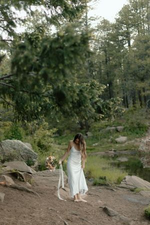 Ethereal wedding scene with flowing white dress against a forest backdrop near a rocky pond.