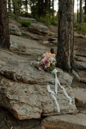 A romantic bouquet with pink and peach flowers rests on a rocky surface with white ribbons.