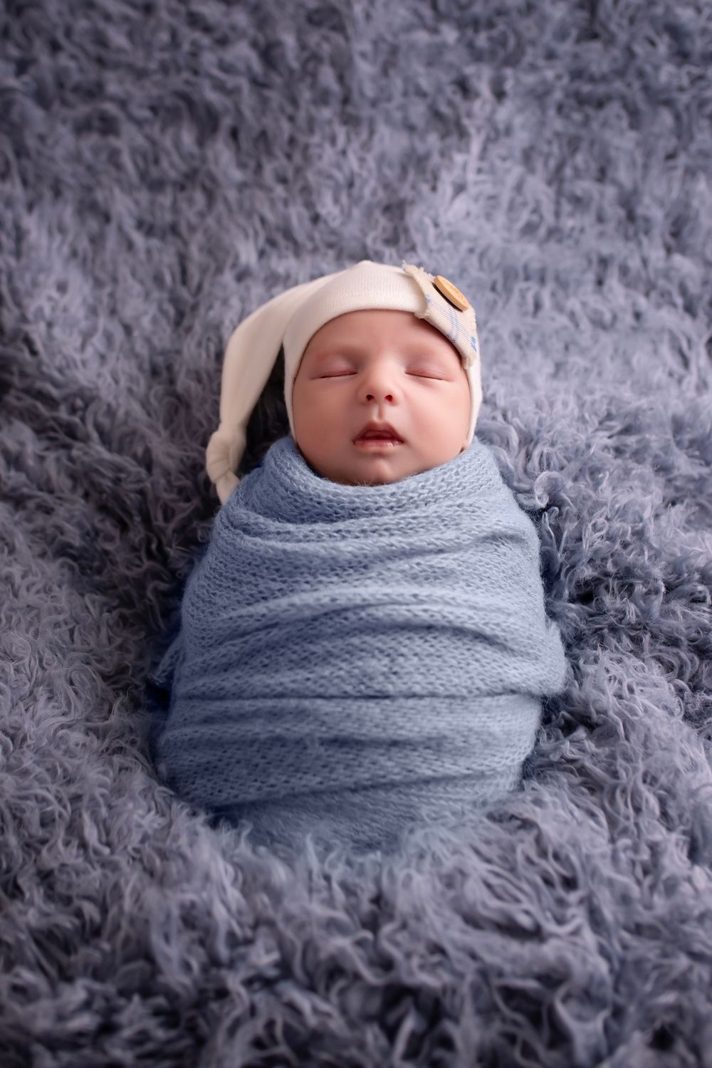 A sleeping newborn swaddled in a soft blue knit wrap rests peacefully on fluffy gray fur.