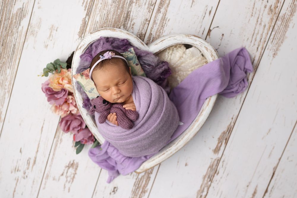 A sleeping newborn in a purple wrap lies on a rustic white wooden backdrop with delicate floral accents.