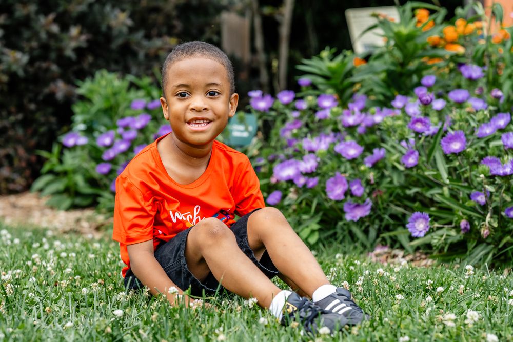 Young child in orange shirt sitting and smiling by purple flowers in a garden.