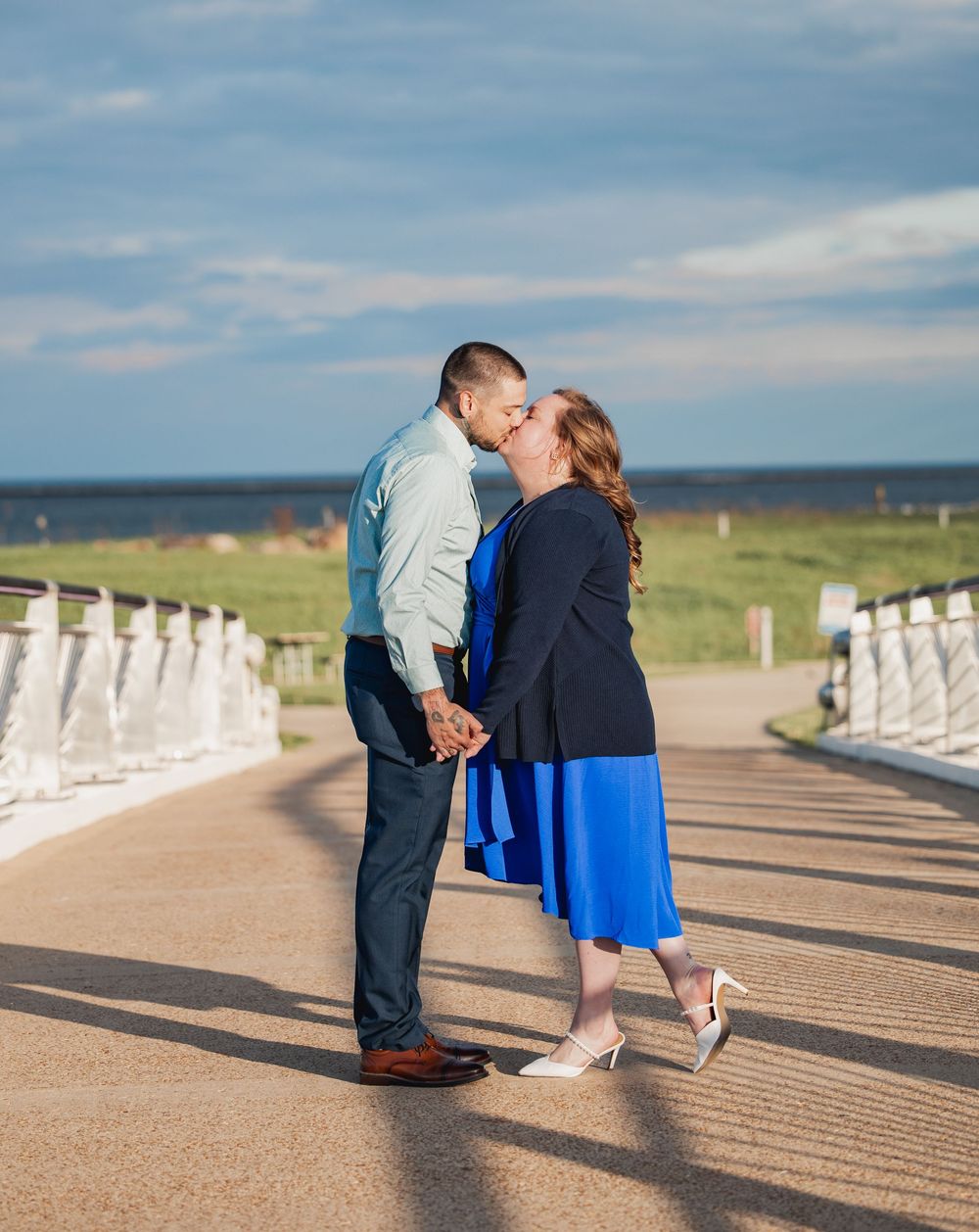 Couple shares a romantic moment on a boardwalk by the ocean with white railing on a sunny afternoon.