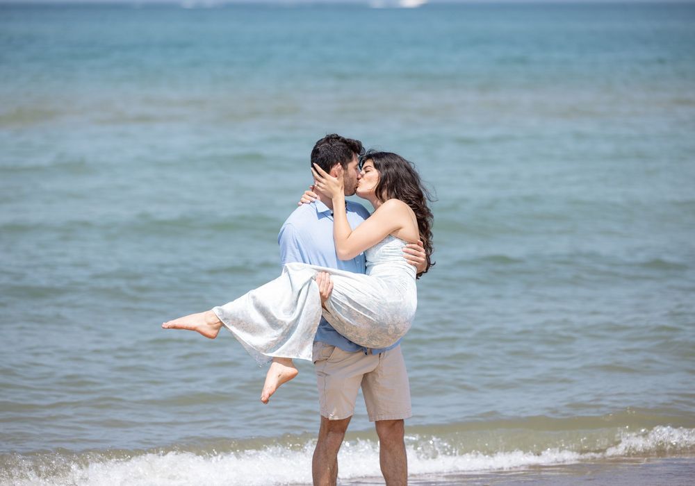 Two people share a romantic embrace on a tropical beach with turquoise waters and white sand in the background.