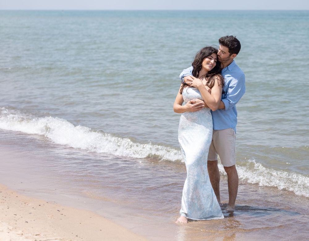 A romantic couple embraces on the sandy beach with gentle waves and blue ocean waters in the background.
