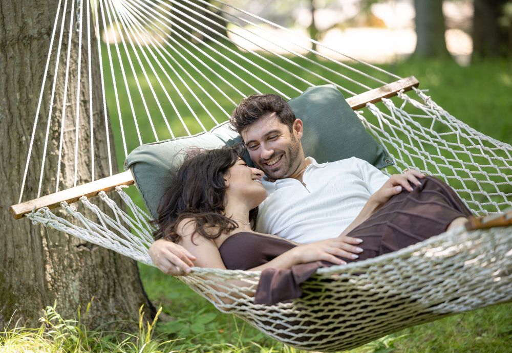 Two people relaxing together and smiling while lounging in a rope hammock outdoors on a sunny day.