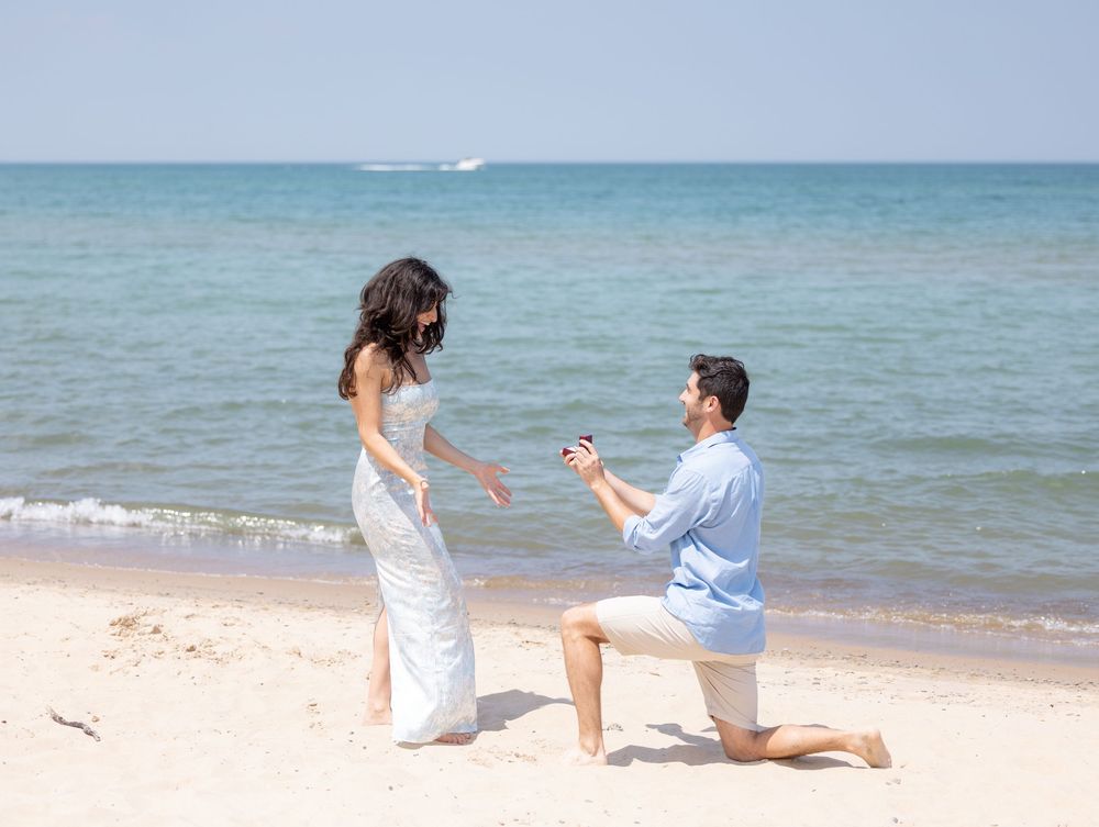 At a sunny beach, two people enact a romantic proposal scene next to the turquoise ocean waters.