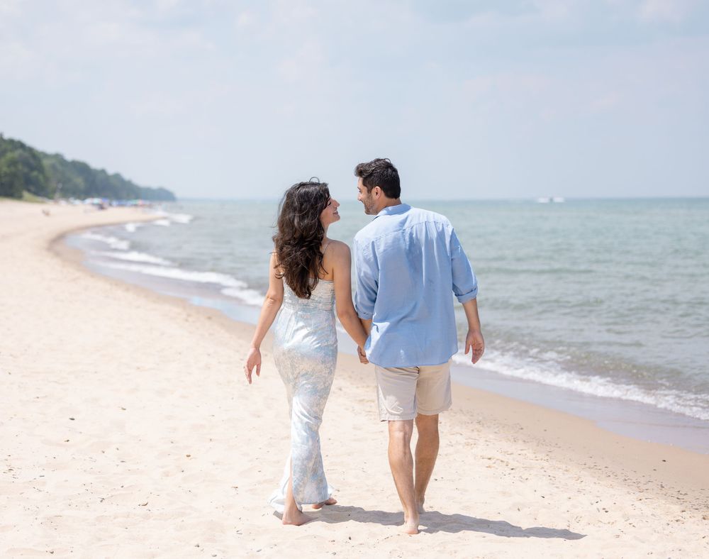 Couple walking barefoot together along a peaceful sandy beach with ocean waves and green hills in the background.