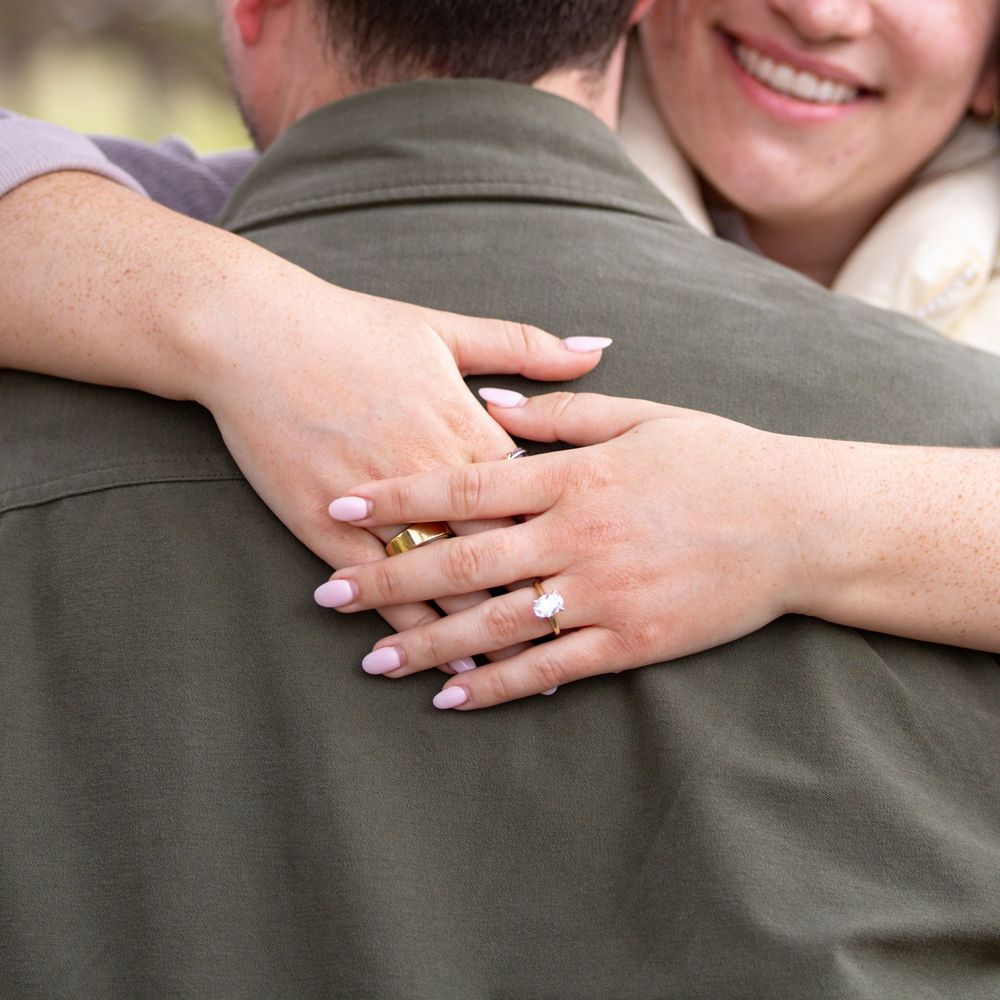 Close up of hands embracing shows engagement ring with freshly manicured nails against grey clothing background.