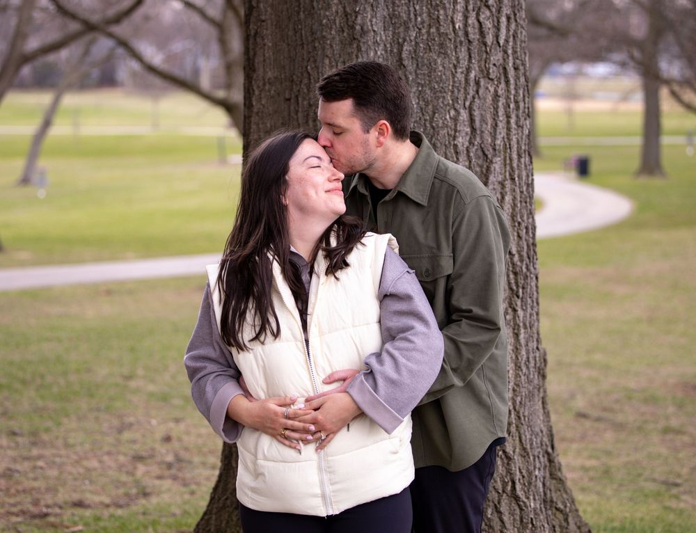 A tender moment between two people sharing a romantic embrace near a tree in a park on a cloudy day.
