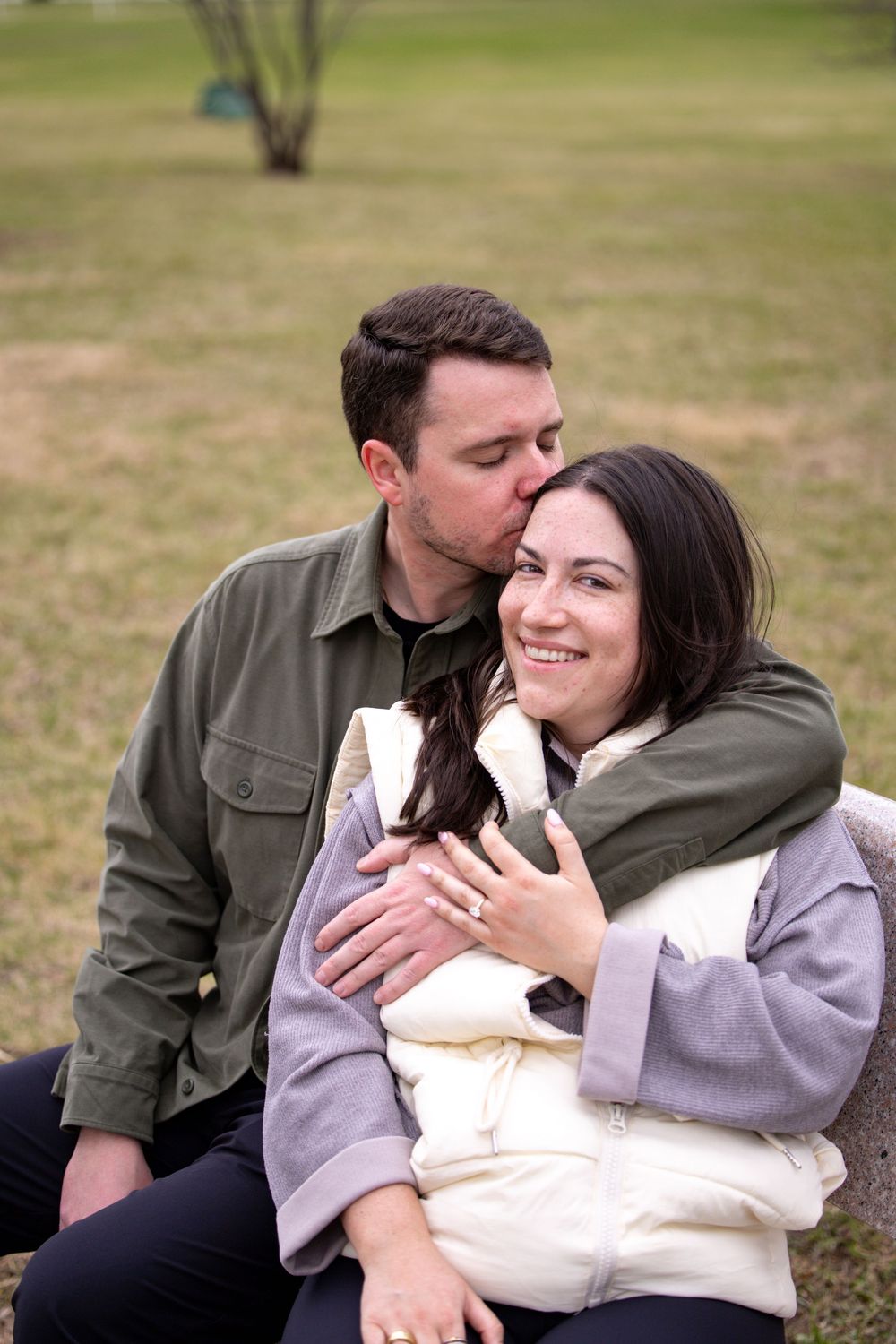 A couple shares an intimate outdoor moment on the grass, embracing and showing affection in casual attire.