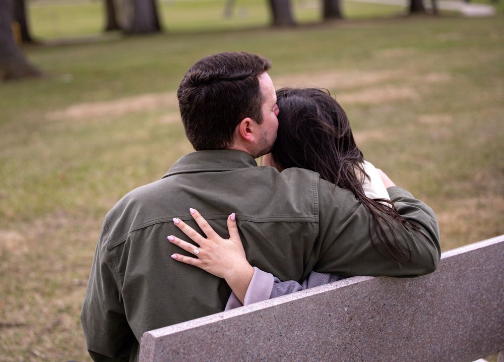 A couple sits close together on a bench in a park setting, sharing an intimate moment while dressed in casual outerwear.
