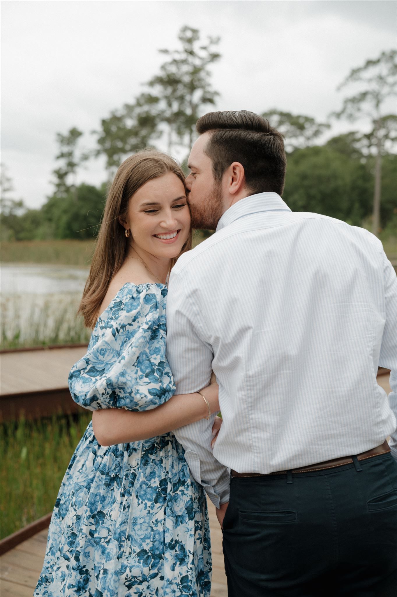 A romantic couple embraces on a wooden dock by a lake, with the woman wearing a blue floral dress on an overcast day.