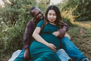 A couple sits together on a blanket in a grassy field during a maternity photoshoot at sunset.