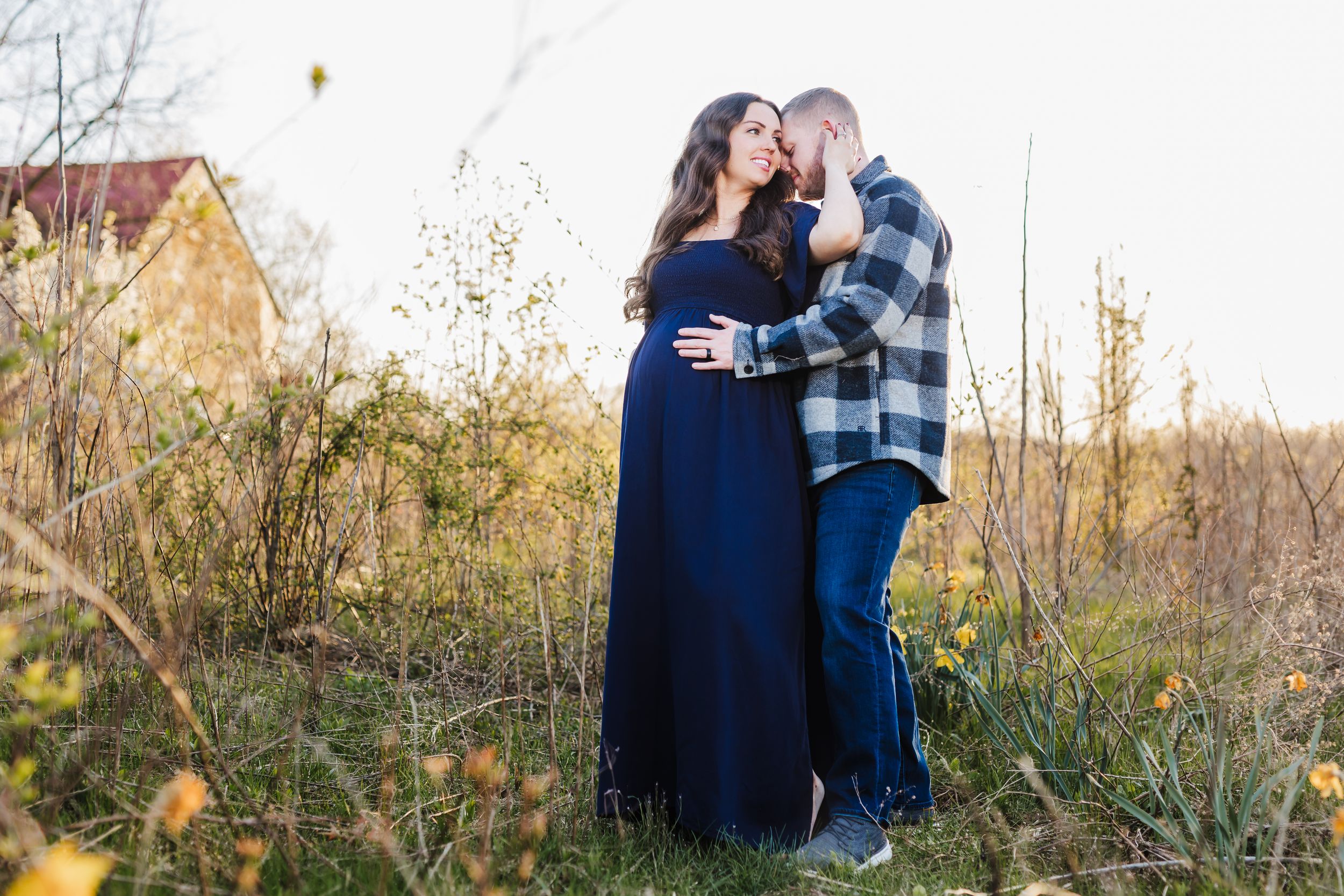 A couple embraces in a rustic outdoor setting with fall foliage, she wearing a long navy dress and he in plaid.