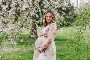 A pregnant woman in a floral dress stands beneath blooming cherry blossoms in a spring garden setting.