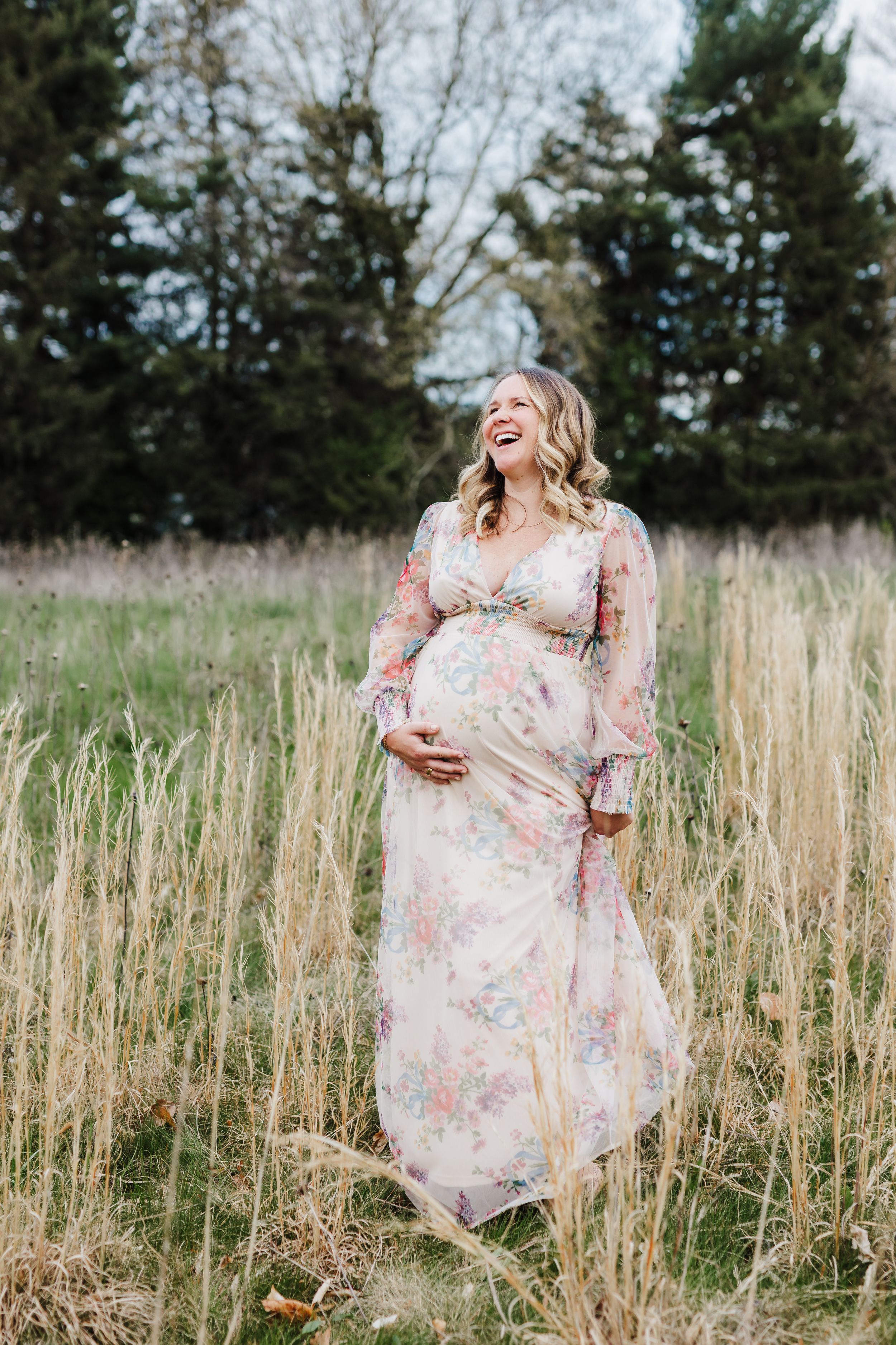 Expectant mother in floral maxi dress laughs joyfully while standing in tall grass field with evergreen trees behind her.