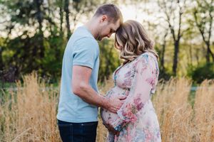 Expecting couple stands together in tall grass meadow during golden hour as they lovingly embrace.