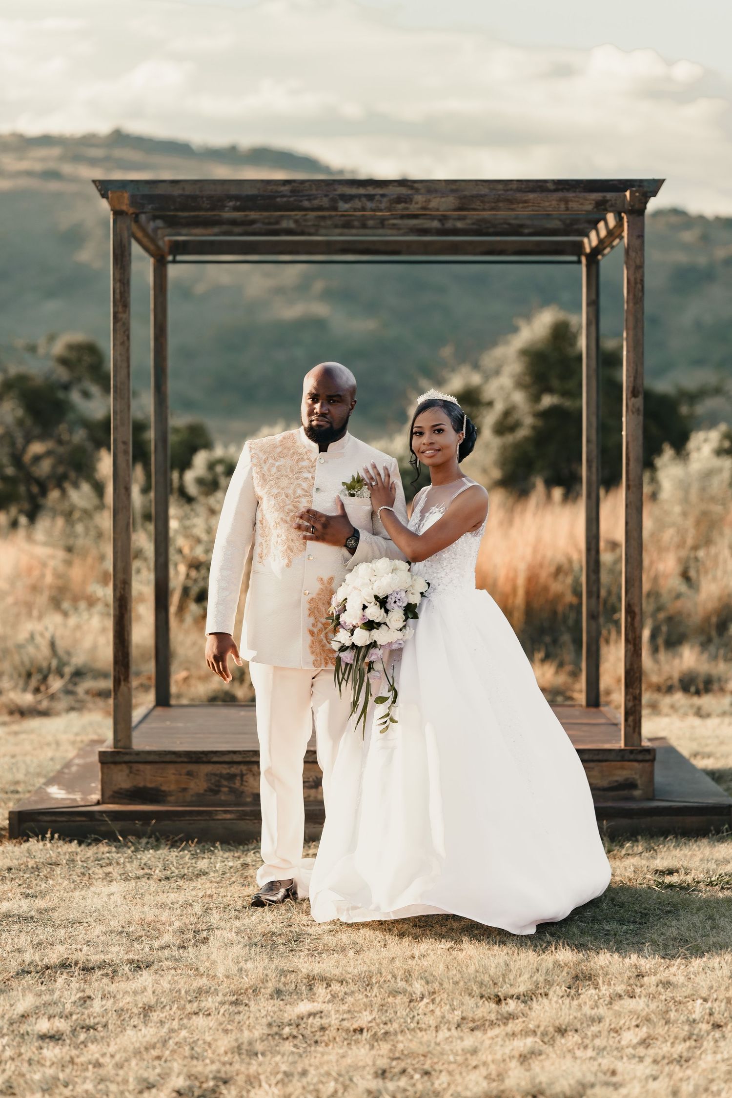 A couple poses in their wedding attire under a wooden arbor in a rustic outdoor setting.
