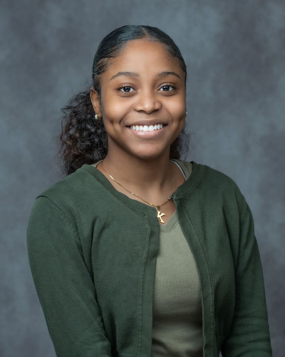 Young woman smiling in green cardigan and gold necklace poses for professional headshot photo.