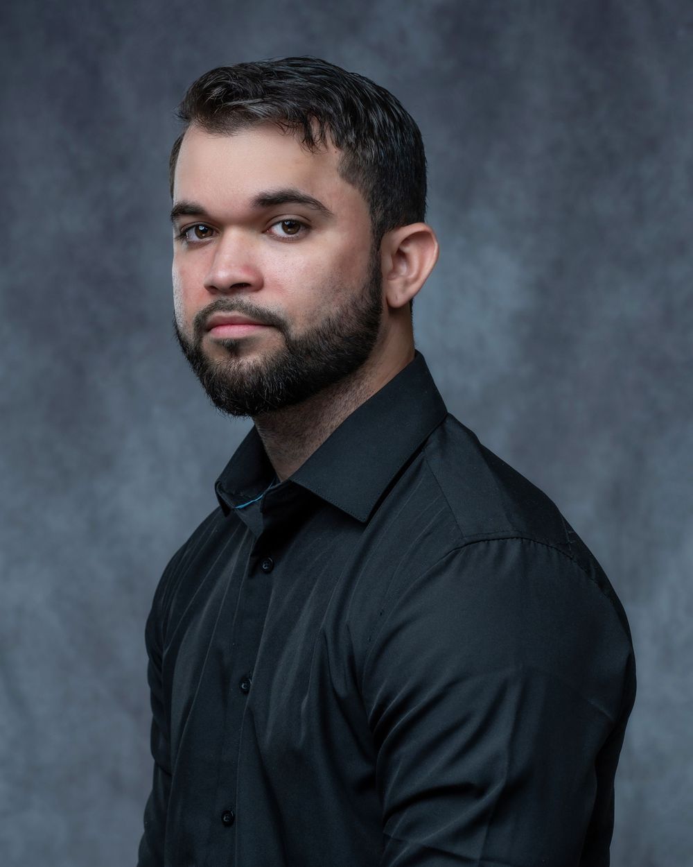 Professional headshot of a young man with a beard wearing a black button-up shirt against a gray backdrop.