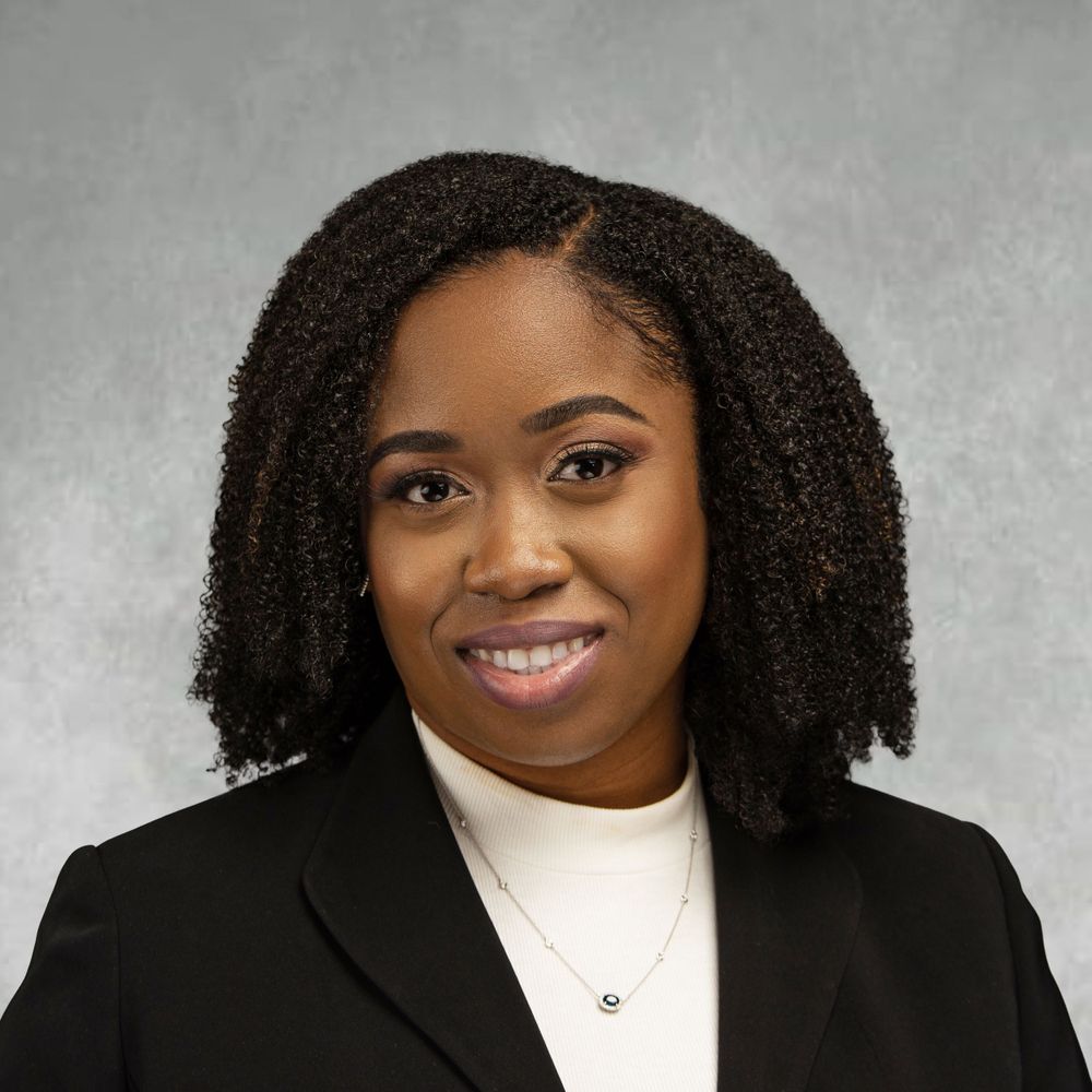 Professional headshot of a young woman in a black blazer and white turtleneck, smiling confidently against a gray background.