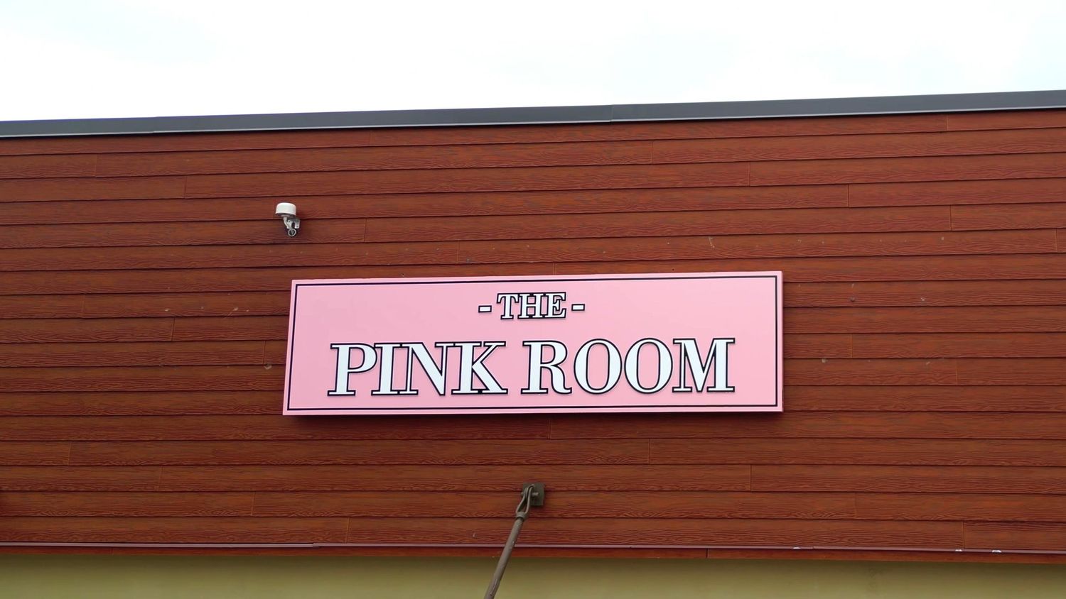 Pink neon sign reading 'The Pink Room' mounted on brown exterior siding.