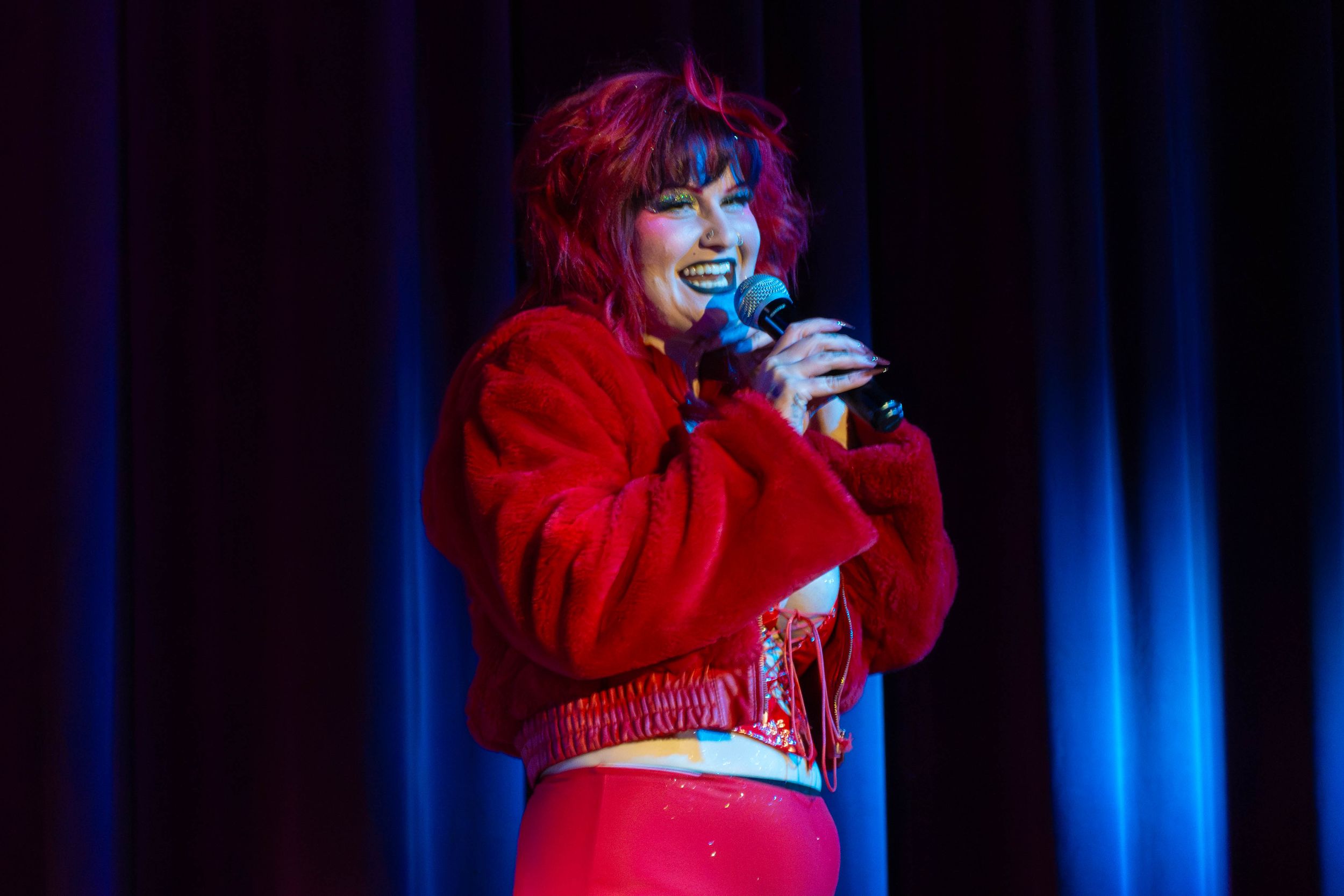 A performer in a bright red outfit sings into a microphone on stage with blue curtains in the background.