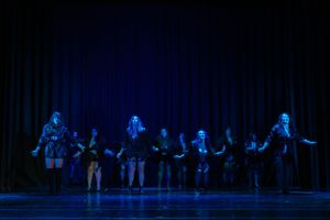 Performers silhouetted in blue stage lighting dance in a line formation during a theatrical performance.