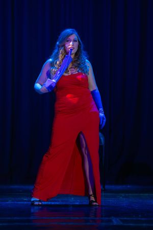 Singer in long red evening gown performs under dramatic blue stage lighting against dark curtain backdrop.