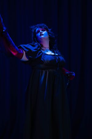 Dramatic stage performer in dark dress illuminated by moody blue lighting against black curtain backdrop.