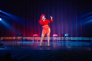 A performer in a red outfit sings on stage under dramatic blue lighting with chairs arranged in the background.